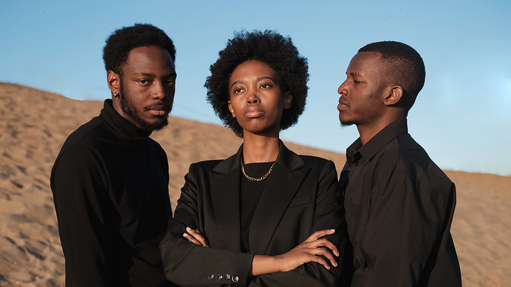 A Black woman stands center with her arms crossed and fixed gaze. A Black man stands to her left staring at her. A Black man on her right stares directly at the camera. All three are dressed in black.