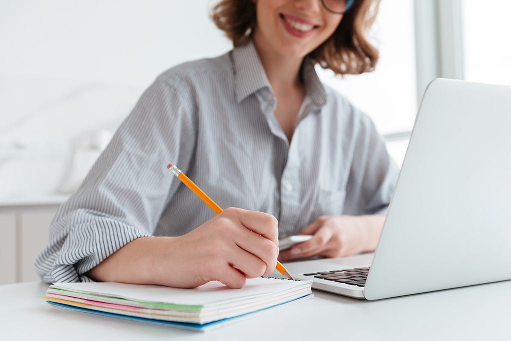 Mulher branca de óculos e cabelo curto, com blusa listrada, sorrindo e tomando nota em caderno, de frente para um notebook aberto.