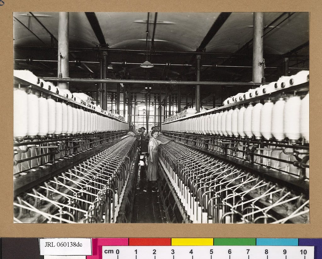 Black-and-white photograph of cotton spinning room featuring young working girls (centre) looking directly at the camera.