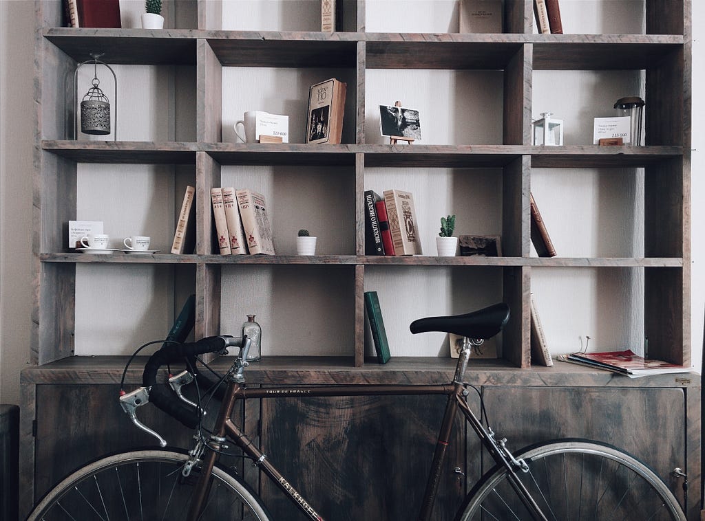 Photo of a gray bookshelf with books sparsely spaced and a road bicycle leaning in front of it. Neutral toned and relaxing to look at.