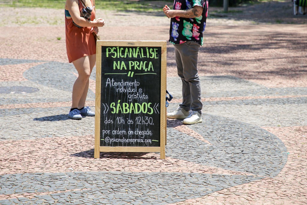 Na praça, de manhã, duas pessoas conversam entre uma placa que anuncia “Psicanálise na Praça: atendimento individual e gratuito aos sábados das 10h às 12h20”. O texto foi escrito com giz, em um quadro negro
