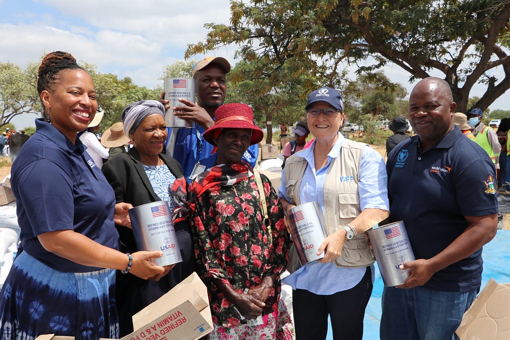 Two older women pose alongside four aid workers holding up cans of USAID-provided food aid. The aid workers are wearing clothing with logos from USAID, WFP, and World Vision.