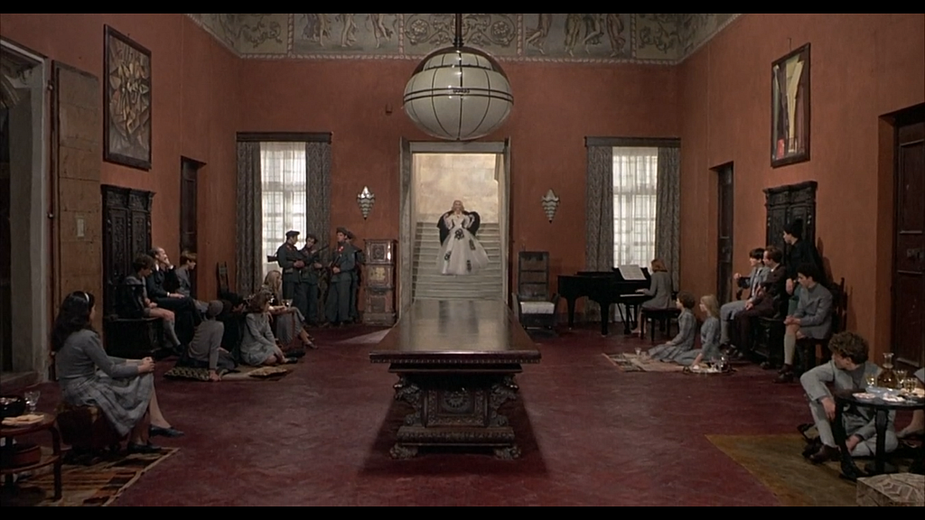 A wide shot of a big, red room, showing the captives sat around the edge and a lot of space between them and a table in the middle. There are paintings on the wall, a low-hanging chandelier, and tall windows. Through an open door, we see Signora Vaccari descending a white white staircase in a white gown and a black shawl. The overall effect is somewhat opulent.