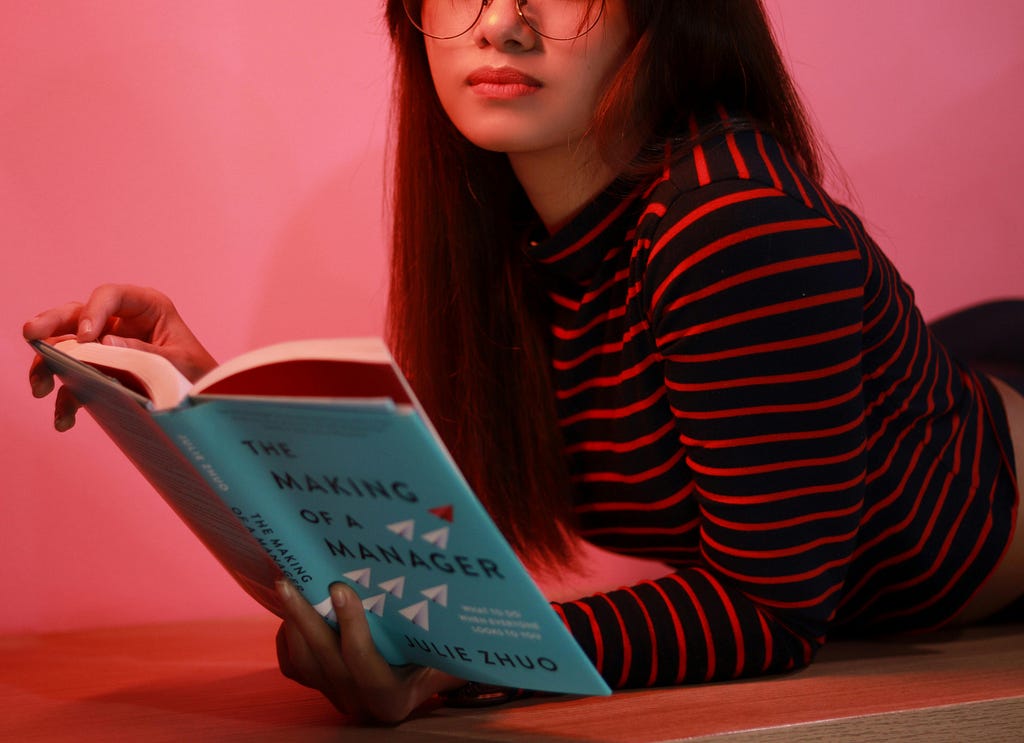 Person wearing a striped red and black top laying on their stomach in a red-tinted room reading a book with a light blue cover
