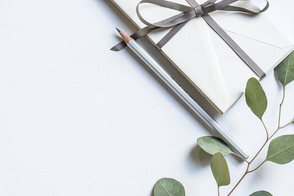 White envelope with a grey ribbon, placed next to a grey pencil and some green leaves.