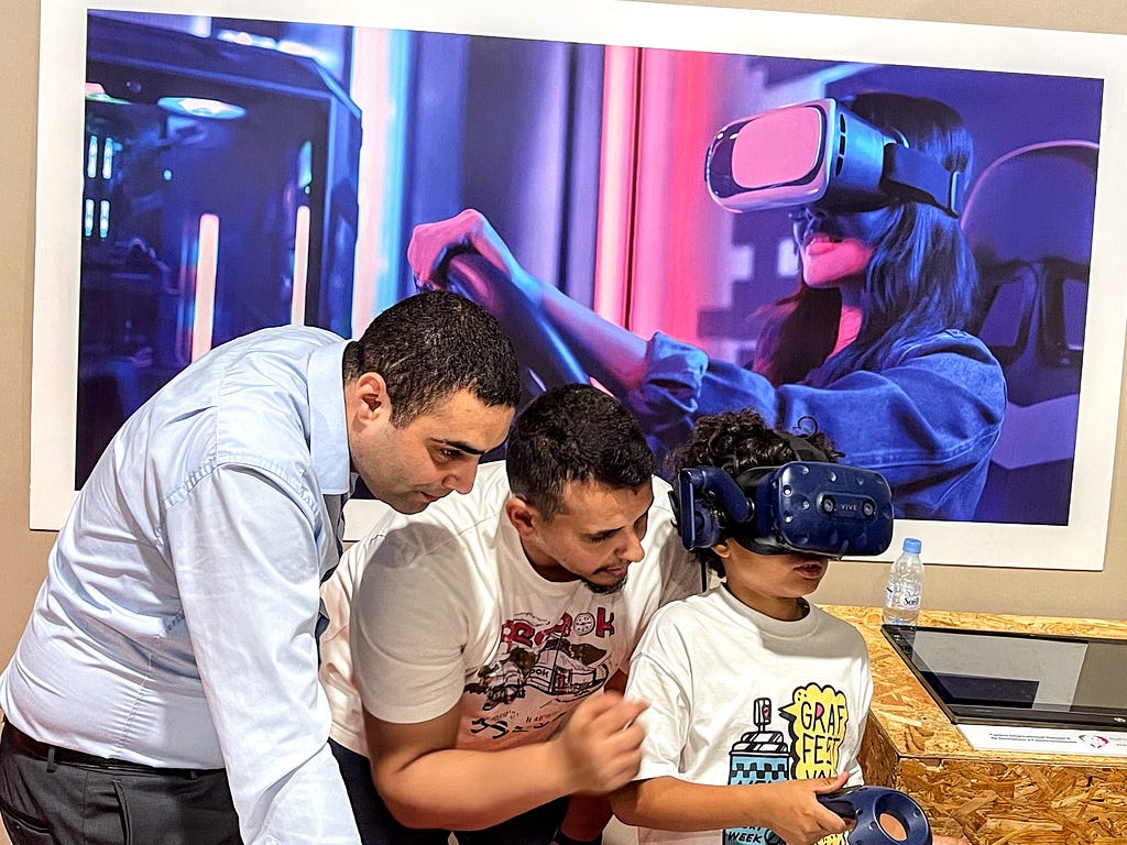 A boy wearing a virtual reality headset and holding a controller is urged on by two men standing behind him. All three stand in front a large photo of a woman wearing a virtual reality headset with her hands on a steering wheel.