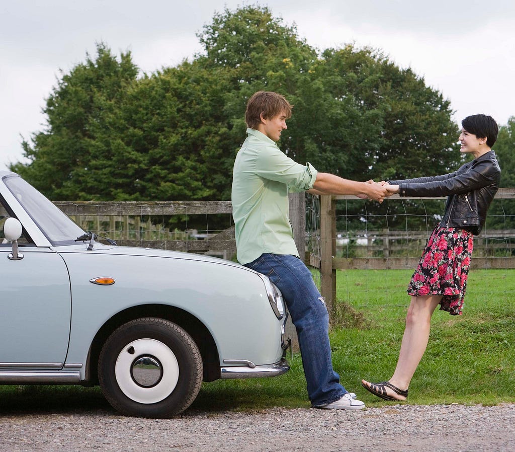 A couple holding hands and balancing, the boy is sitting on a car, the girl trusts the boy to not let go