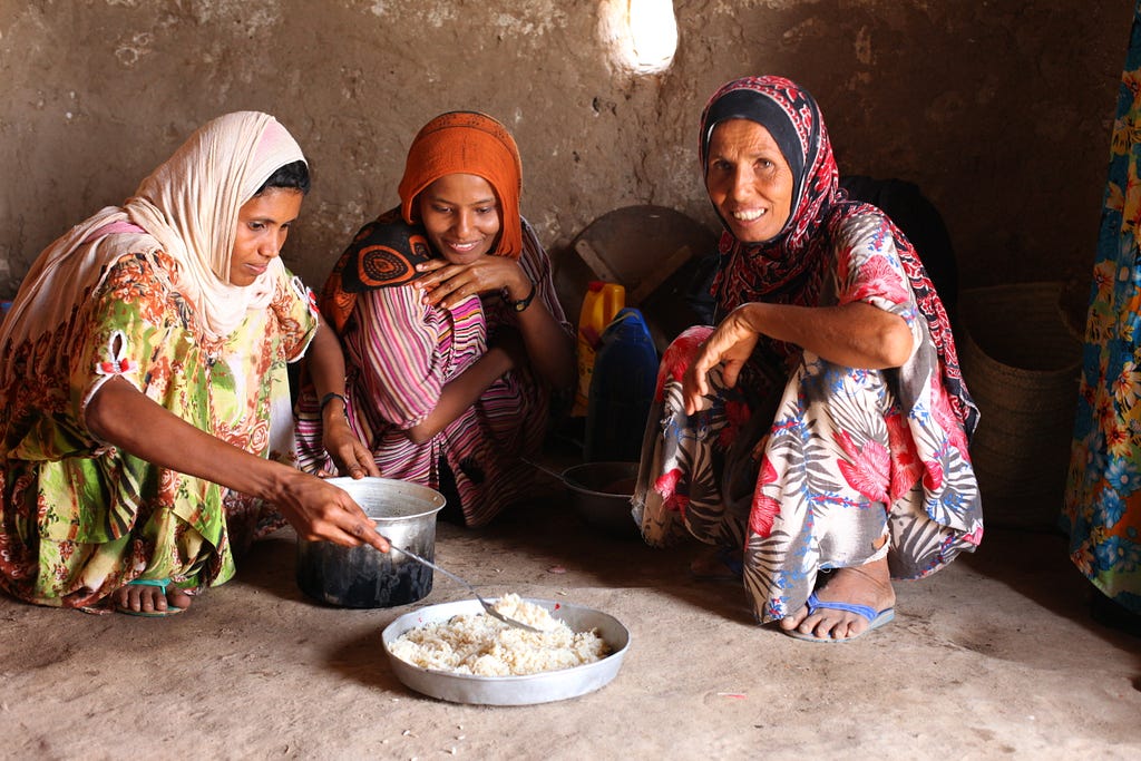 Three woman sitting in on the floor; one of them is serving some rice on a plate of food in front of them.
