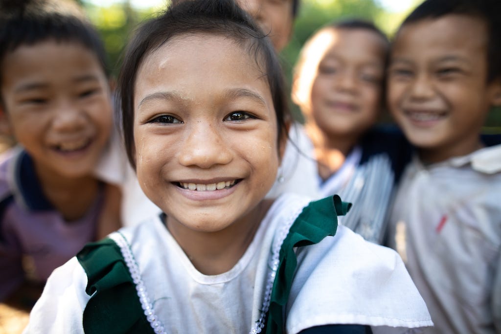 Tarchanee, a young Thai girl, is smiling and standing outside wearing a green and white shirt. Her friends are behind her.