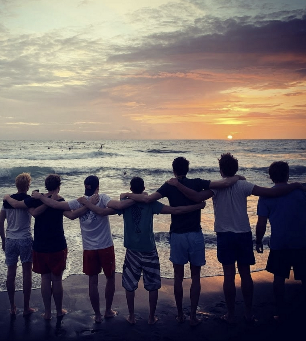 Image Description: 7 of the Ava Team members standing on a beach, holding each others shoulders watching the sunset with the water in the background.