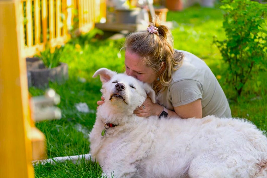 Woman kissing a white dog that’s laying down on its forehead.