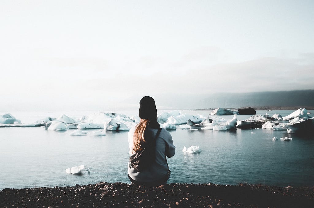 A girl sitting with her back to us on a black beach in front of water filled with iceberg pieces. A moody setting.