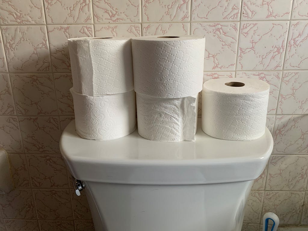 The back of a white toilet with five toilet paper rolls sitting on it. There is pink and white marble tile behind the toilet.