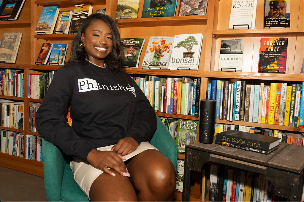 Photo of Black woman in a library sitting next to a table that has a black coffee tumbler with &lsquo;Dr. Iriah&rsquo; on it. On the table there are also two books stacked on top of each other: one that says Dr. Sade Iriah on top and one under it that says &lsquo;The addicted brain&rsquo;. She is smiling ear-to-ear and is wearing a Black crewneck that says Ph.inisheD. in white and black letters. She has wavy black hair with brown highlights. There are a bunch of different books in her background.