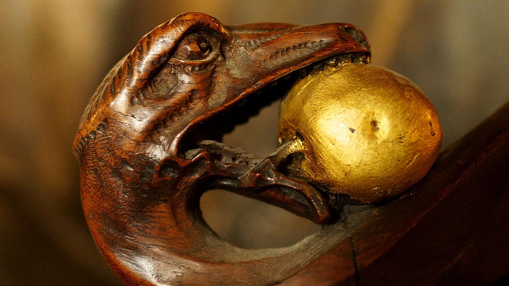 Finely detailed wood carving of a snake with human eyes, holding in its mouth, as if in presentation, a gold-colored fruit