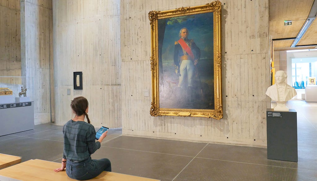 A young girl, seen from behind, sits on a bench and looks at a work of art displayed on a concrete wall in a museum. She holds a tablet in her hand. The work represents a man who seems to be a military man with a red scarf and white pants.
