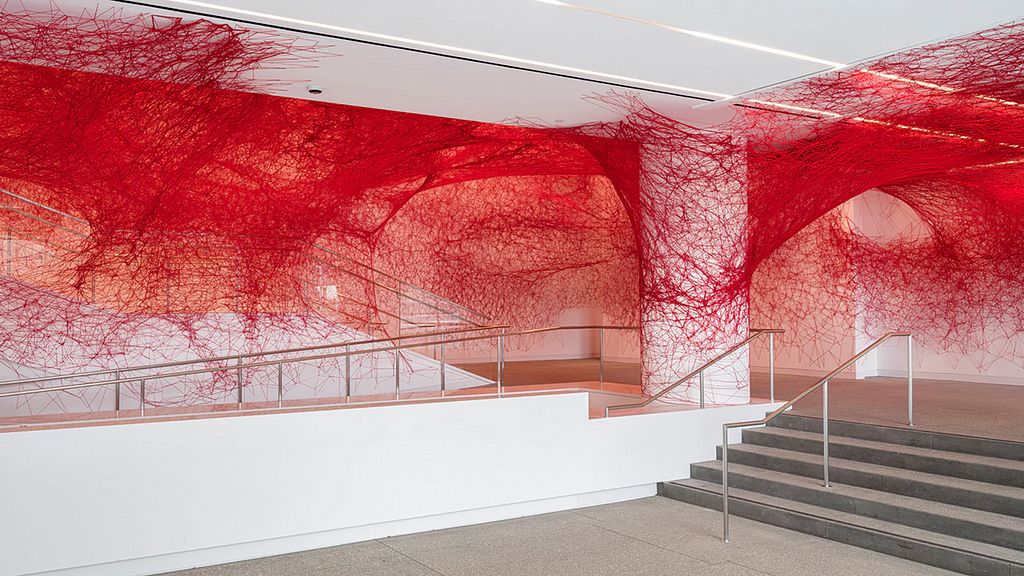 Standard white walls and concrete floor of a modern museum lobby. Stairs are to the right and there’s a central column. Along the walls, ceiling, and even the column is red yarn, like a massive, messy spiderweb.
