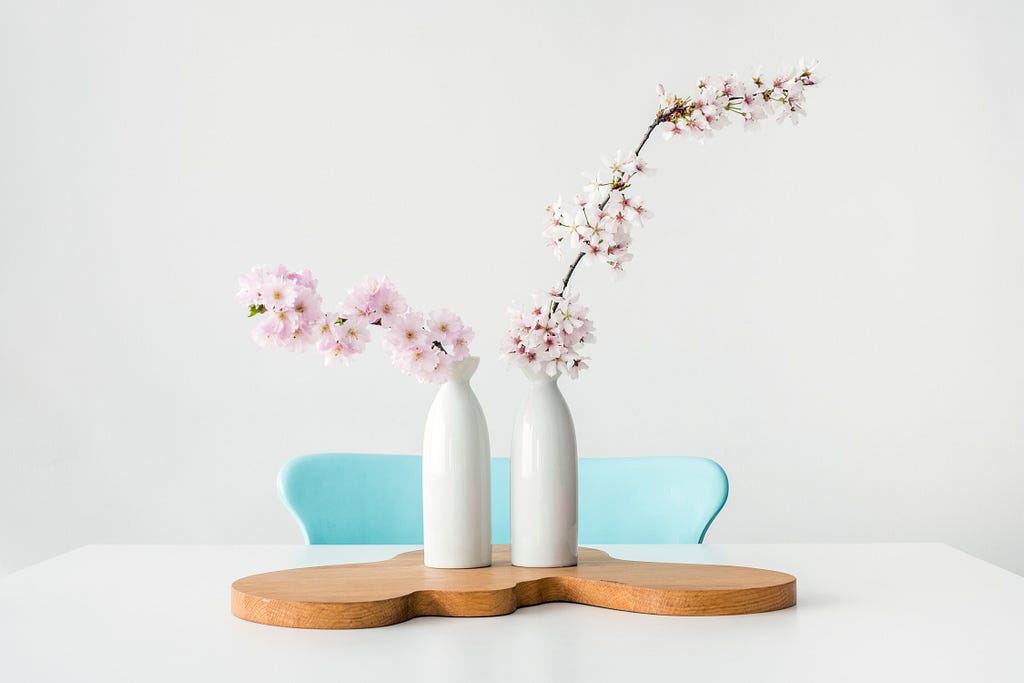 Two stems of blossom in white vases on a white table with a blue chair