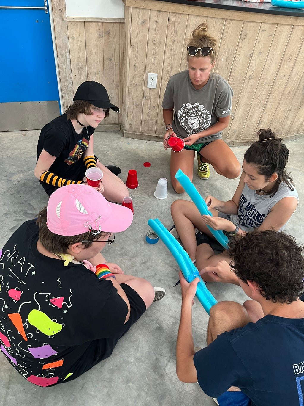 campers and activity leader have various supplies on the ground including pool noodles, tape, and cups. Th are discussing their strategy for creating a marble tunnel.