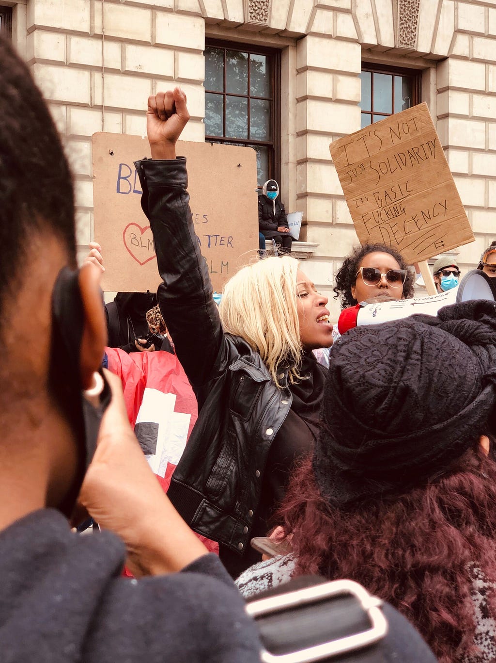 Black woman speaking into a megaphone at a protest and a sign in the back that says “…it’s basic fucking decency”