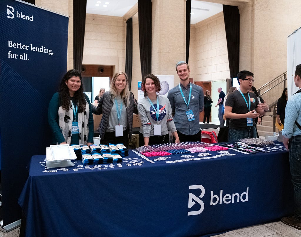 The Blend talent team stands, smiling, behind their branded career fair table, covered with swag, at Tech Inclusion 2019