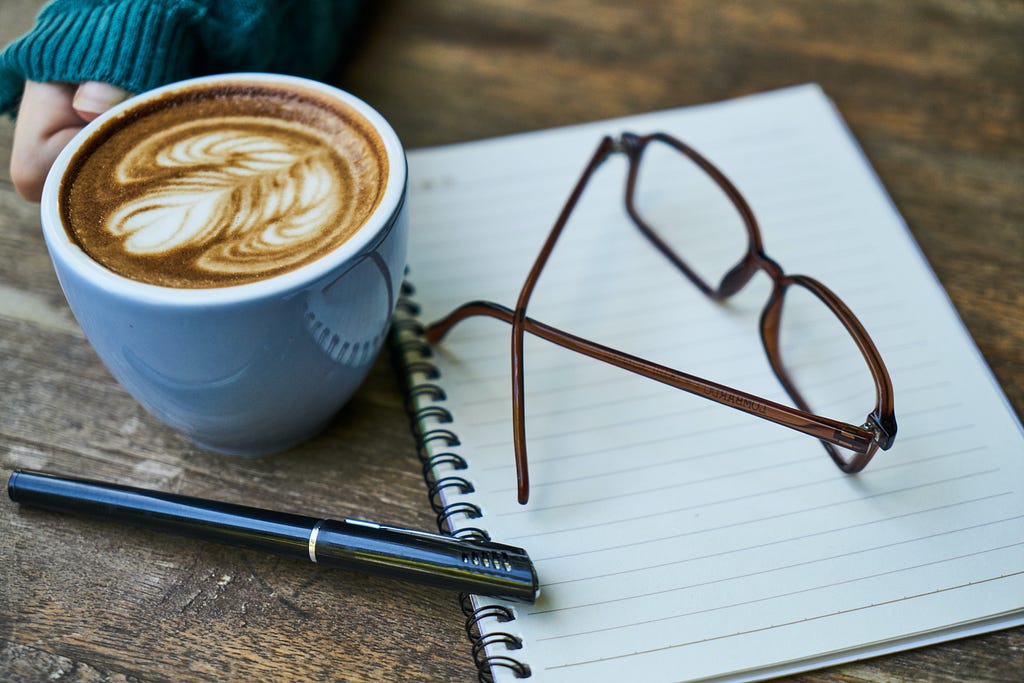 a hand holding a cup of coffee near lying on the table, brown glasses, notebook and a pen