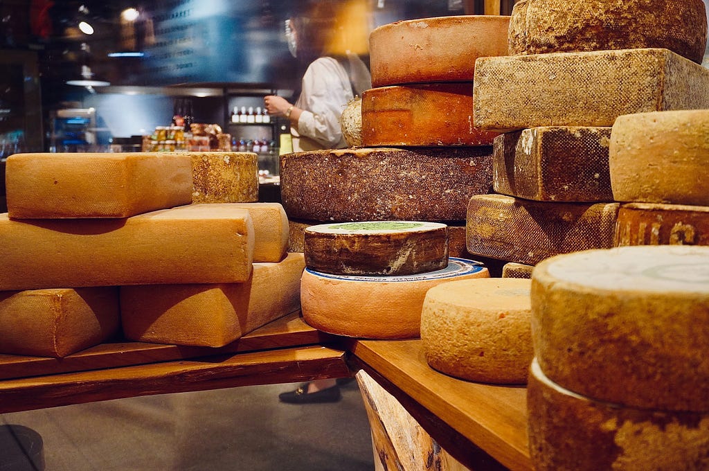 A pile of different types of cheese in a shop. A lady walks past in the background.