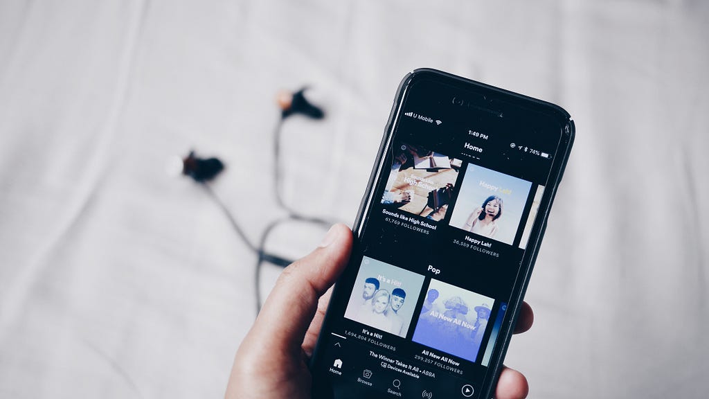 A hand with a smartphone inside where the Spotify app is opened. In the background earphones are lying on a sheet.