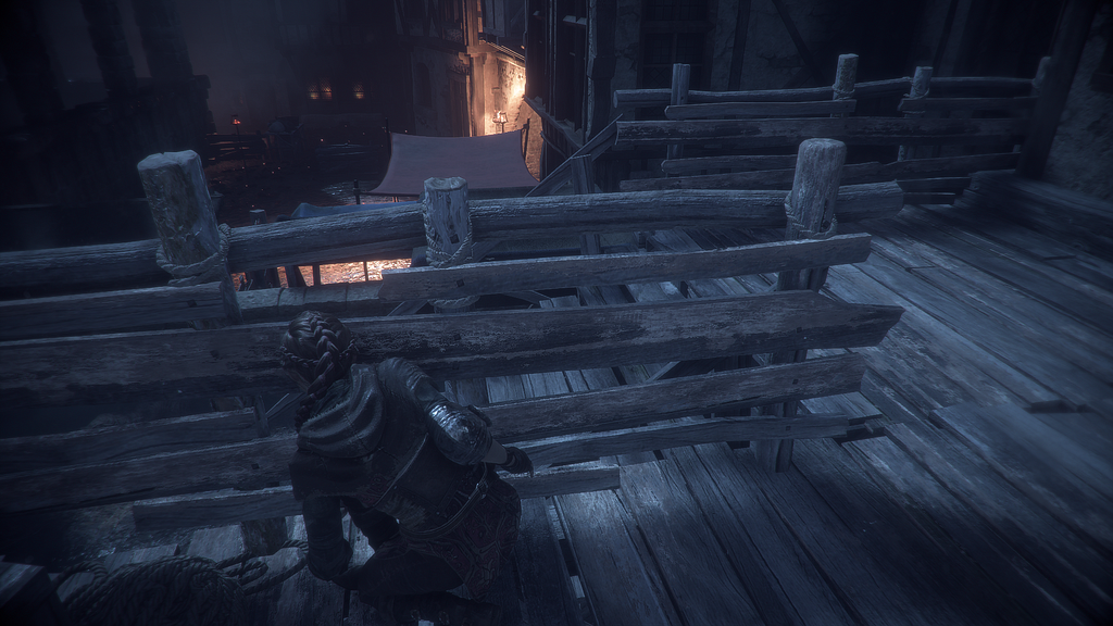 A screenshot from A Plague Tale: Innocence, taken at night. In the foreground, a young girl with her hair in a braid is crouched behind a wooden railing on top of a set of stairs. Below her is what looks like a village square with a few fires and torches in places.