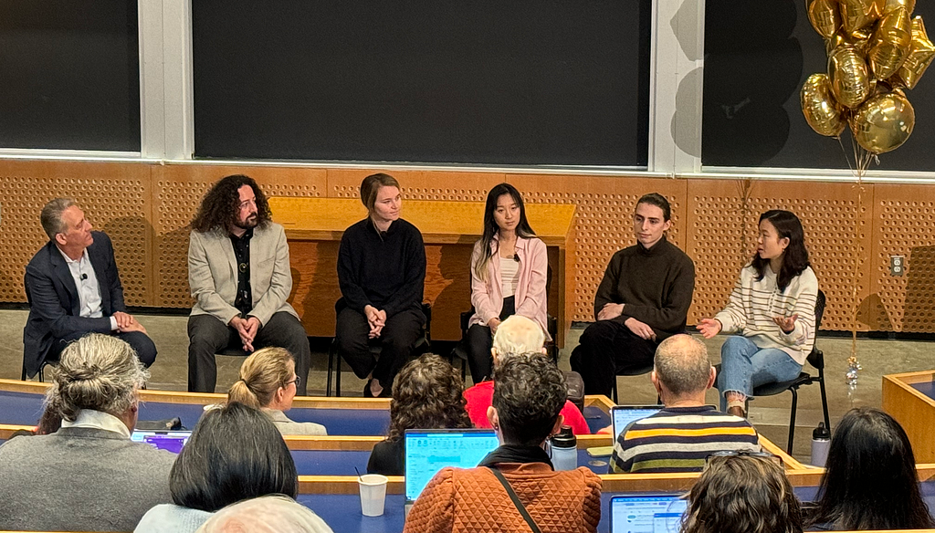 Candid photo of six adults seated in the front of a college classroom with three rows of a dozen or more adults sitting in the foreground.