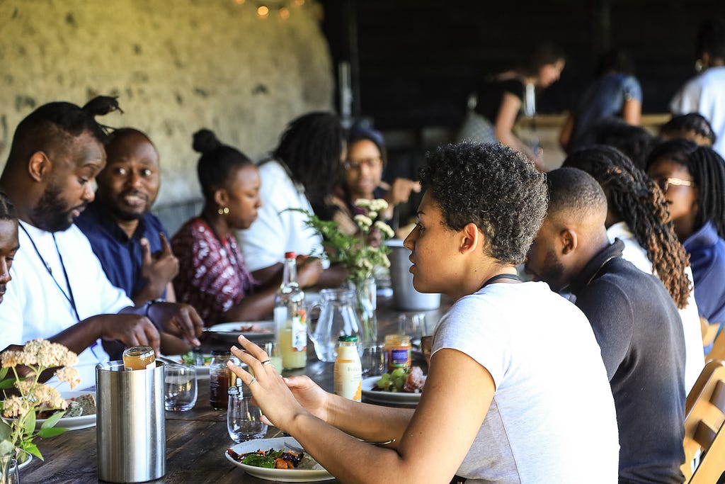 Multiple guests sit around a long trestle table with drinks, food and flowers on it.