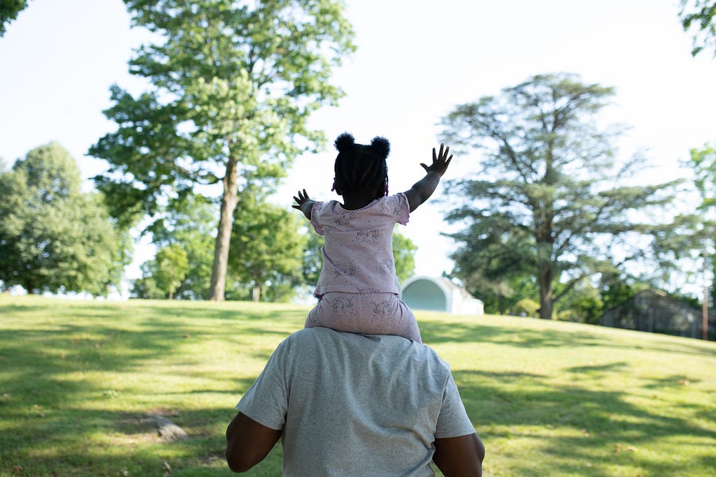 An adult is carrying a small child on their shoulders. The child is reaching out excitedly with both hands toward a park-like landscape.