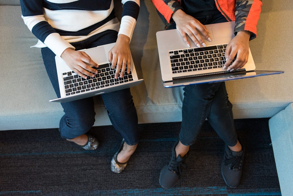 Photo from above of two women of color sitting on a couch, typing on macs.