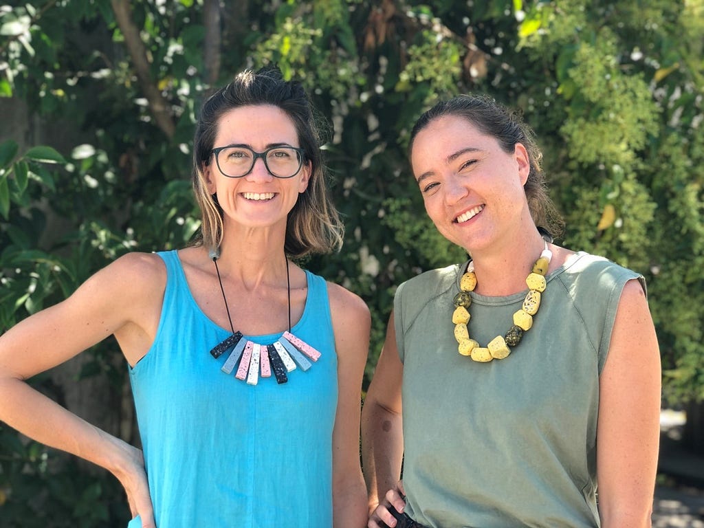 Two woman standing next to each other. One is wearing a light blue tank top, one is wearing a light green one. They are both wearing homemade necklaces of colorful rocks.