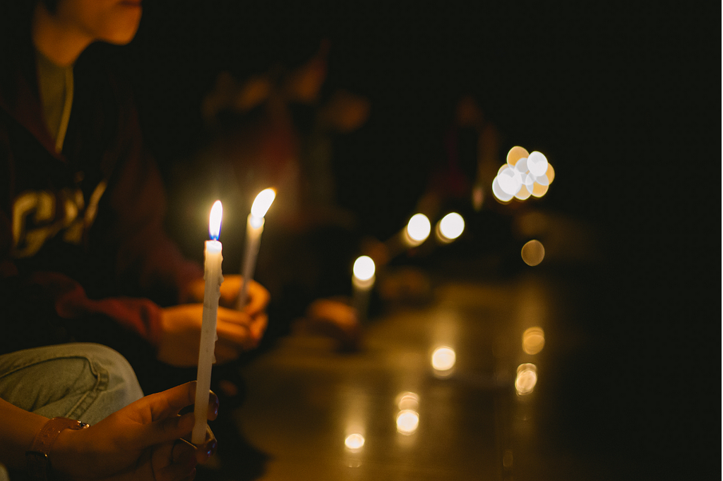 A hand holding a candle with a flame, with another person holding a lit candle also sitting in the background.