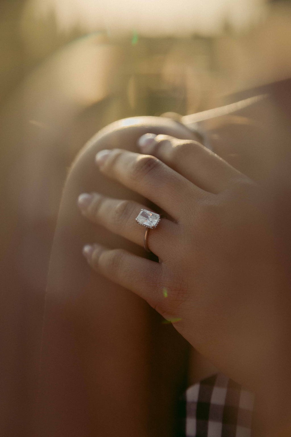 Photo of a diamond ring on a persons hand, which is resting on their shoulder. The picture is taken outside at dusk as the sun is fading.