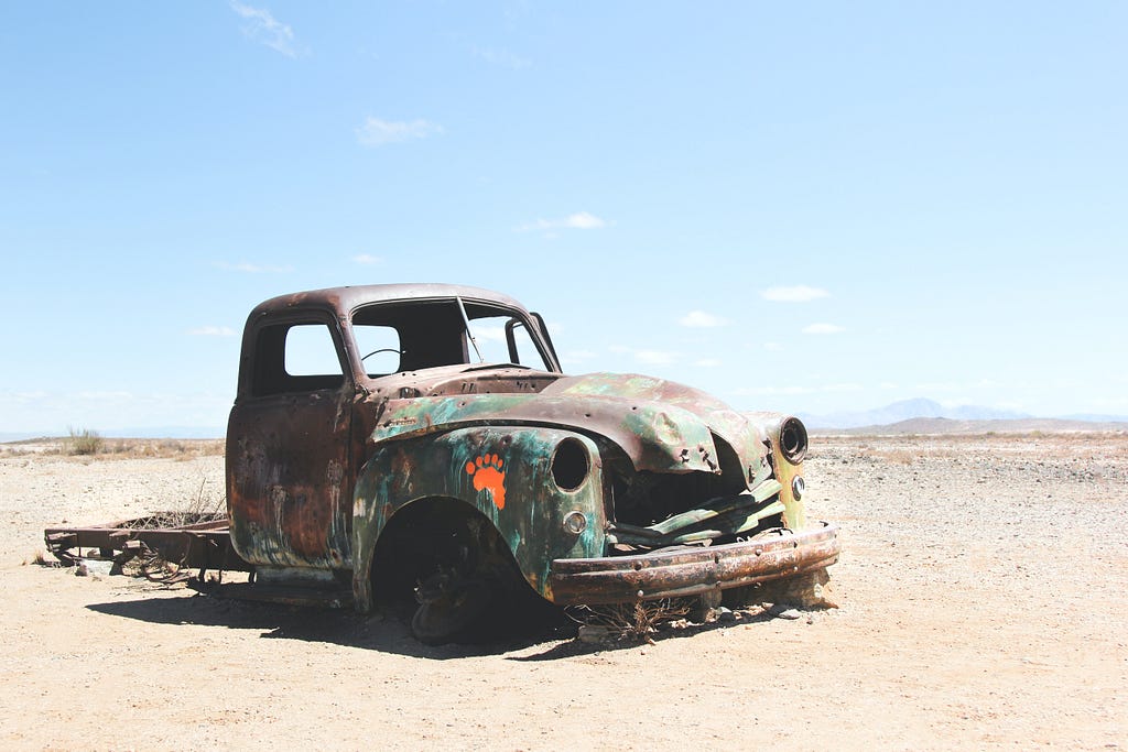 An abandoned truck, with no tires, no windows, and no back part, in the middle of a desert.