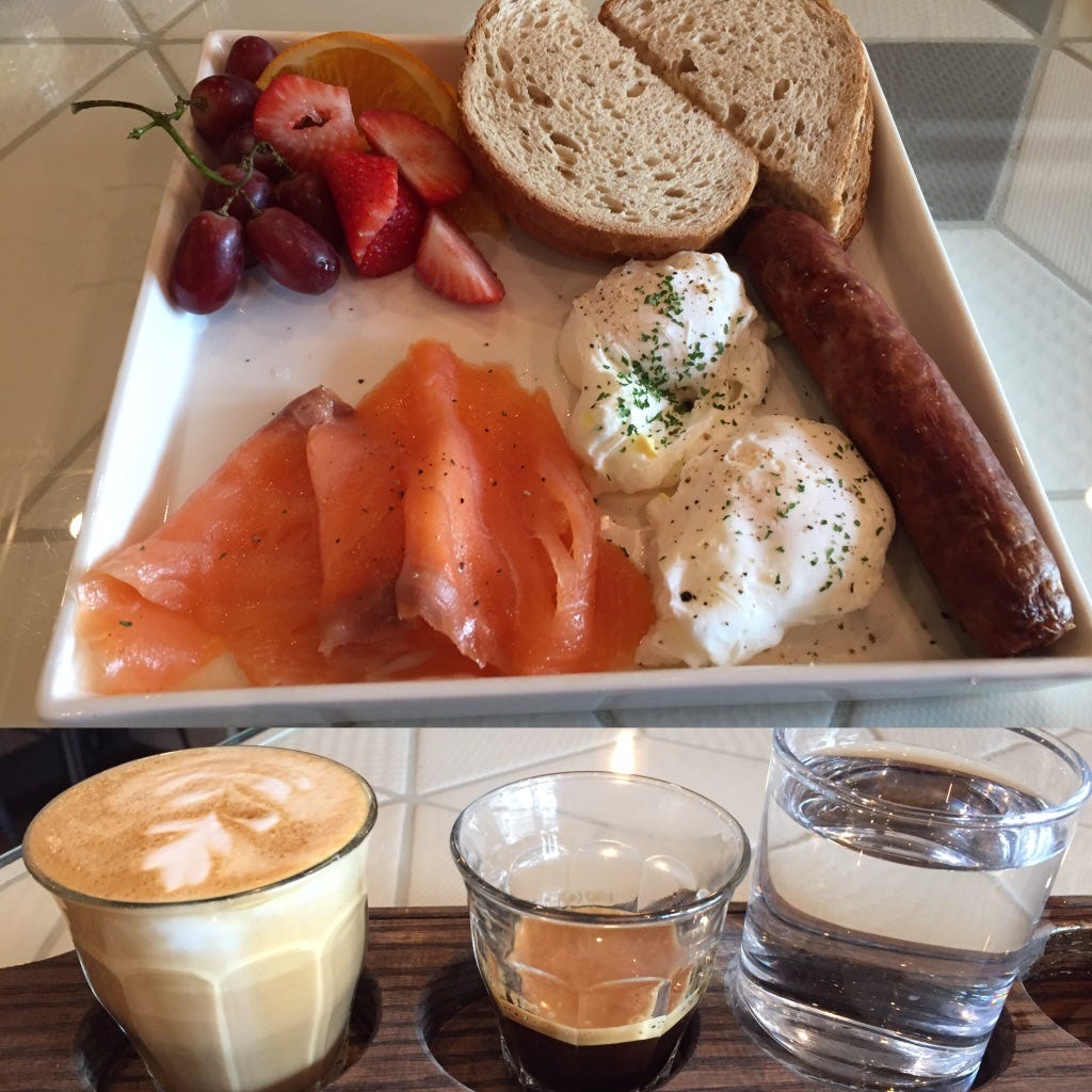 Smoked salmon, eggs, sausage and toast with two glasses of coffee and a glass of water in the foreground.