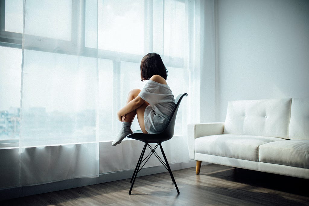 A woman sitting on a chair with her legs to her chest looking out the window.