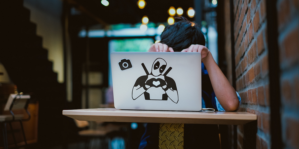 Image of a person working behind their laptop that features a camera sticker and a decal of Deadpool.