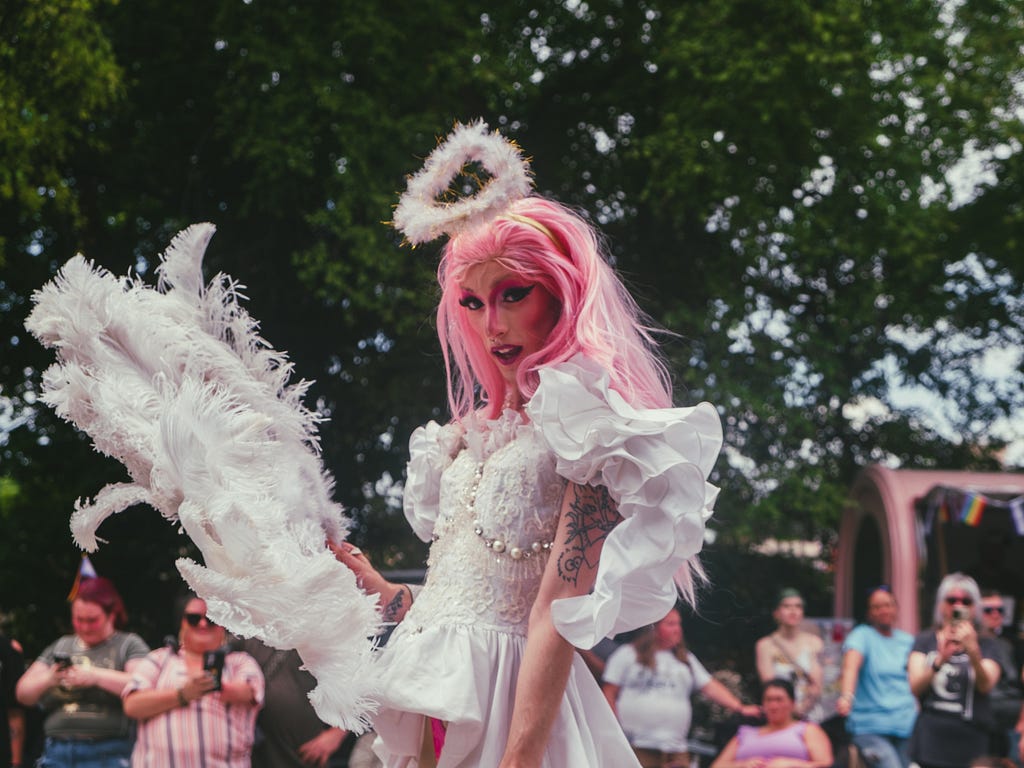 A drag queen in a white, ruffly dress & wearing a white, fuzzy halo. They have a bright pink wig & a large, white, feathered fan