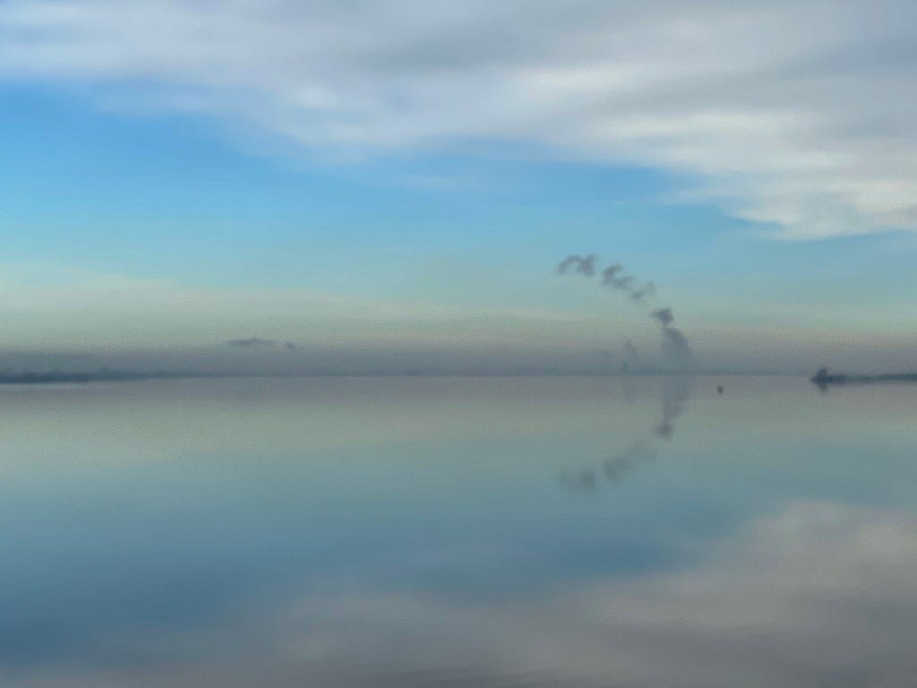 View over still water with blue sky and clouds. A bank of fog sits on the horizon with a plume of smoke arcing up and reflected in the water from distant industrial buildings
