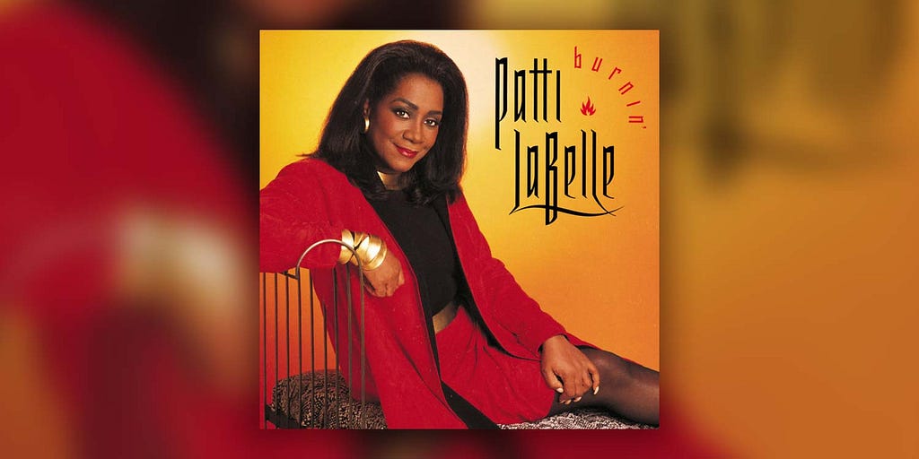 Patti LaBelle’s ‘Burnin’’ album cover: she sits on a chaise lounge with a backing made of thin, curved metal railing. She wears a red skirt suit, with a black top, gold bangles on her right wrist, a gold geometric necklace, and a shiny, gold belt.