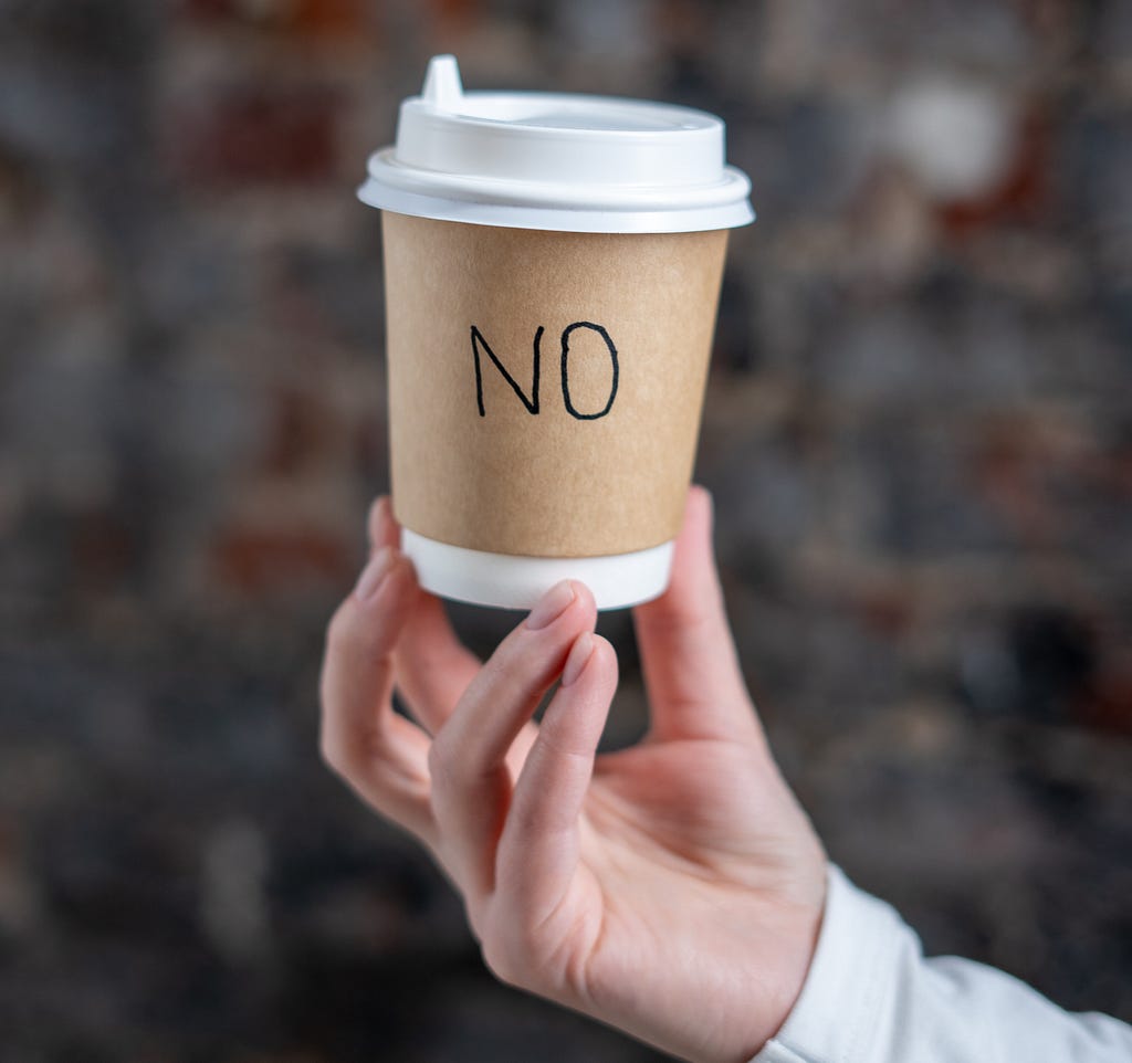 A hand is holding up a disposable coffee cup with the word “NO” written on it.