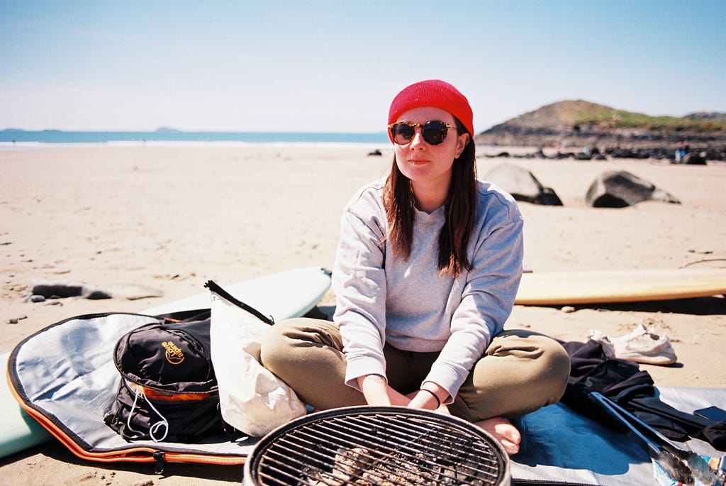 A person sitting in front a bbq, wearing a grey jumper, sunglass and a red hat. In the background is a beach and sea on the horizon.
