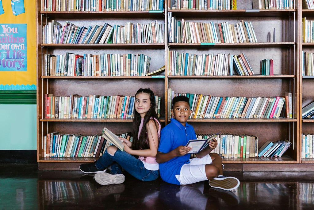 Two school-age children sit on the floor in front of tall bookshelves filled with books. A partial view of a blue and yellow poster can be seen to the left of the bookshelves. The children are sitting back-to-back and they are each holding a book and looking sideways into the camera.