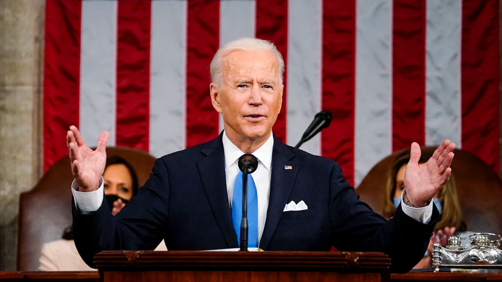 President Joe Biden addresses Congress from a podium. His hands are raised and he is wearing a black suit with a blue tie. Behind him sit Vice President Kamala Harris and Speaker of the House Nancy Pelosi. The American flag can be seen in the background.