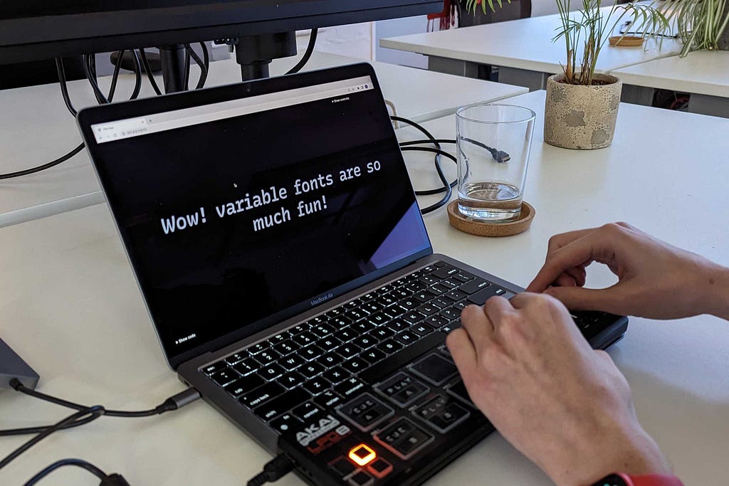 A laptop with the text, “Wow! variable fonts are so much fun!” on the screen. In front of it is a long rectangular device with large buttons and dials. Two hands are twiddling the knobs.