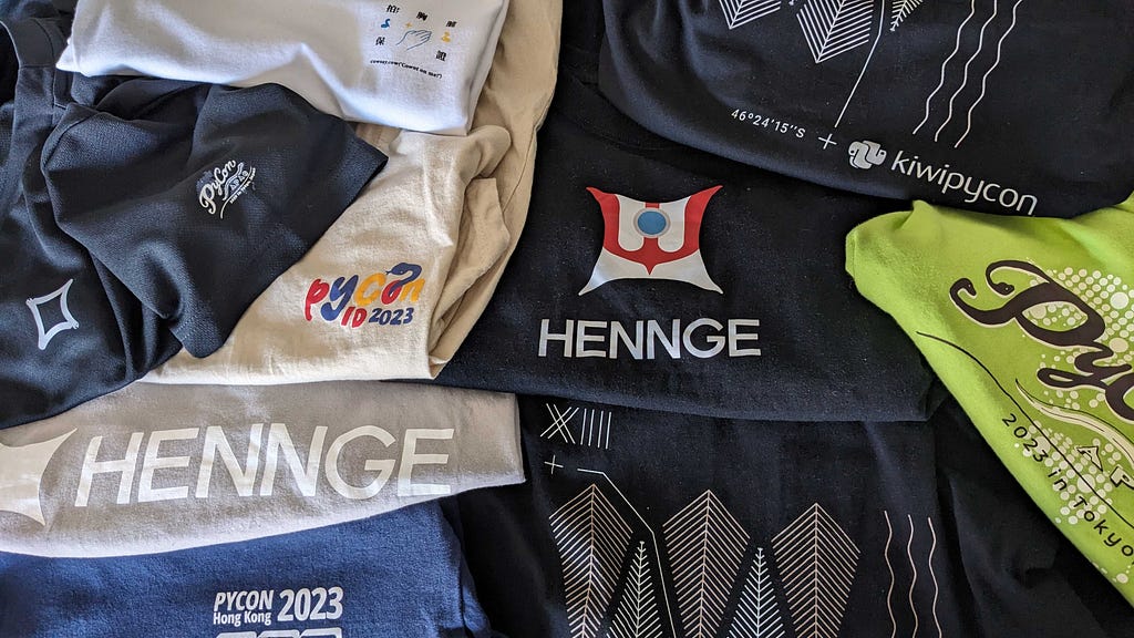 A pile of multicolored t-shirts with several PyCon and HENNGE logos visible.