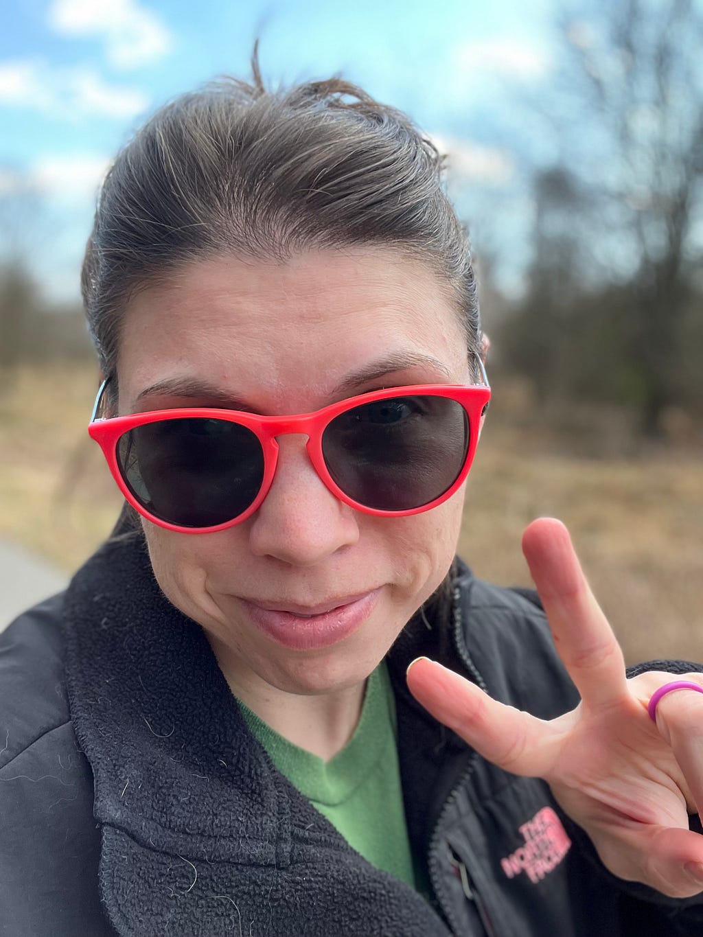 Bri wearing orange sunglasses, on a walking trail in the woods, smiling at the camera and giving the peace sign.